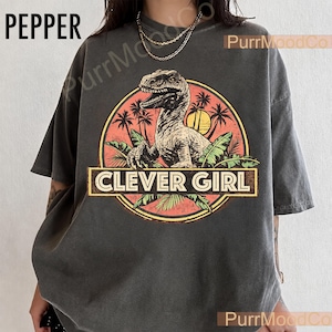 Può includere: Una t-shirt grigio antracite a maniche corte con una grafica circolare raffigurante un dinosauro, palme e un sole giallo. Il testo CLEVER GIRL è stampato in un banner rettangolare al centro del design. La t-shirt ha un girocollo.
