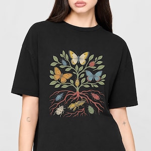 Op de afbeelding: Een zwart t-shirt met een centrale afbeelding van een boom met zichtbare wortels. De takken van de boom bevatten diverse kleurrijke vlinders en insecten, terwijl het wortelstelsel omringd is door extra kevers en insecten in aardetinten.