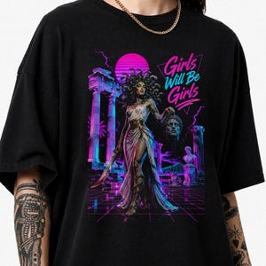 Peut inclure: Un t-shirt noir avec une impression graphique vibrante représentant Méduse tenant une tête coupée au milieu de ruines grecques antiques. Le design utilise des couleurs rose et bleu néon, incluant un coucher de soleil, des éclairs et le texte Girls Will Be Girls.