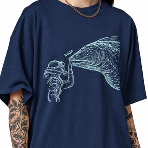 Może przedstawiać: Granatowy t-shirt z jasnoniebieską grafiką przedstawiającą nurka w hełmie w stylu vintage, dotykającego nosa dużej mureny. Napis BOOP jest nadrukowany jasnoniebieskim kolorem nad palcem nurka, który dotyka pyska ryby.