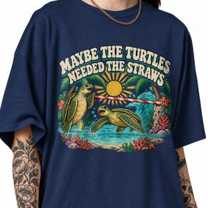 May include: A navy blue t-shirt featuring a graphic of two green sea turtles in the ocean with palm trees, a yellow sun, and coral. The text MAYBE THE TURTLES NEEDED THE STRAWS is printed above the turtles, who are holding a red and white striped straw.