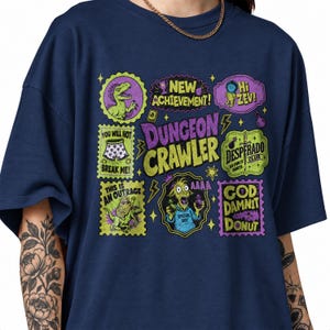 Könnte beinhalten: Ein marineblaues T-Shirt mit einem Grafikdruck, der in der Mitte den Text "Dungeon Crawler" zeigt. Um den Text herum befinden sich verschiedene bunte, briefmarkenartige Illustrationen, darunter ein Dinosaurier, eine Ziege und Phrasen wie "New Achievement!" und "God Damnit Donut."