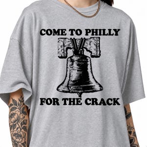 Op de afbeelding: Een grijs gemêleerd t-shirt met een zwarte afbeelding van de Liberty Bell met een duidelijke barst in het midden. De tekst "COME TO PHILLY" is in het zwart boven de klok gedrukt en de tekst "FOR THE CRACK" staat in het zwart onder de klok.