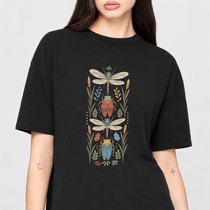 Peut inclure: Un t-shirt noir avec une impression graphique verticale représentant deux libellules et deux cigales disposées en colonne. Les insectes sont entourés d'éléments botaniques stylisés, dont des massettes et des feuilles aux tons sourds de rouge, bleu et jaune.