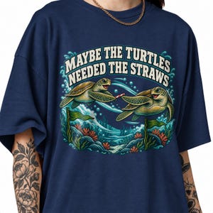 Peut inclure: Un t-shirt bleu marine avec une illustration de deux tortues de mer vertes nageant sous l'eau. Une tortue tient une paille rayée rouge et blanche. Le texte au-dessus des tortues indique "MAYBE THE TURTLES NEEDED THE STRAWS" en lettres blanches.