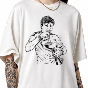 Könnte beinhalten: Ein cremefarbenes T-Shirt mit kurzen Ärmeln und einer schwarzen grafischen Illustration einer Person, die Spaghetti direkt aus einem großen Pokal isst. Die Person hat lockiges Haar und trägt ein T-Shirt mit Streifen an den Schultern.