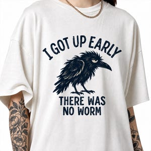 Op de afbeelding: Een off-white t-shirt met korte mouwen en een donkerblauwe afbeelding van een norse kraai. De tekst boven de kraai luidt "I GOT UP EARLY" en de tekst onder de kraai luidt "THERE WAS NO WORM" in een vetgedrukt, donkerblauw schreefloos lettertype.