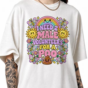 Könnte beinhalten: Ein cremefarbenes T-Shirt mit einer bunten Grafik, die einen lächelnden Regenbogen, Sonnenblumen und Schmetterlinge zeigt. Der Text lautet I NEED 2 MALE VOLUNTEERS FOR A POO, ergänzt durch ein braunes, lächelndes Kackhaufen-Emoji.