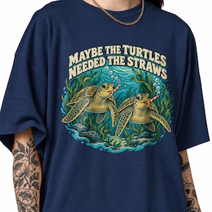 Könnte beinhalten: Ein marineblaues T-Shirt mit einer grafischen Illustration von zwei Meeresschildkröten, die unter Wasser zwischen Seetang und Felsen schwimmen. Jede Schildkröte hat einen rot-weiß gestreiften Strohhalm im Mund. Der Text MAYBE THE TURTLES NEEDED THE STRAWS ist in einer cremefarbenen, gebogenen Schrift über den Schildkröten gedruckt.