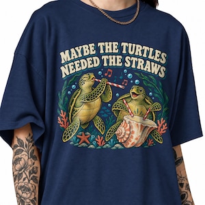 Könnte beinhalten: Ein marineblaues T-Shirt mit einer Grafik von zwei Meeresschildkröten und rot-weiß gestreiften Strohhalmen. Eine Schildkröte trinkt aus einer großen Muschel, die andere hält einen Strohhalm. Der Text lautet "MAYBE THE TURTLES NEEDED THE STRAWS".
