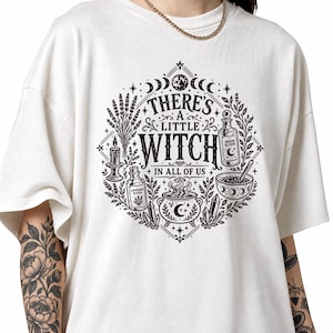 Può includere: Una t-shirt color bianco sporco con una stampa grafica nera di un design circolare con il testo "THERE'S A LITTLE WITCH IN ALL OF US". L'illustrazione include un calderone, cristalli, flaconi "MOON ELIXIR" e "LAVENDER ELIXIR", una candela e lavanda.