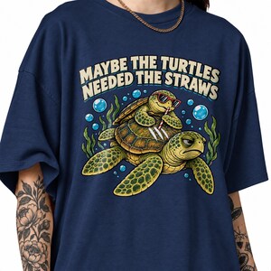 Könnte beinhalten: Ein marineblaues T-Shirt mit einer Grafik von zwei grünen Meeresschildkröten unter Wasser. Eine Schildkröte trägt eine rote Sonnenbrille und benutzt einen Strohhalm, während die andere zusieht. Darüber steht der Text "MAYBE THE TURTLES NEEDED THE STRAWS".