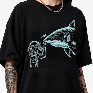Könnte beinhalten: Ein schwarzes T-Shirt mit kurzen Ärmeln und einem hellblauen Grafikdruck, der einen Taucher zeigt, der die Nase eines großen Hais berührt. Der Text BOOP ist in Hellblau über dem Finger des Tauchers gedruckt, der die Schnauze berührt.