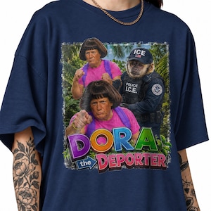 May include: A navy blue t-shirt featuring a graphic print of two figures with brown bob haircuts wearing pink shirts and purple backpacks, alongside a monkey in a dark blue police uniform with an ICE cap, with the text DORA the DEPORTER.