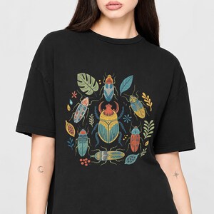 Op de afbeelding: Een zwart T-shirt met korte mouwen en een centrale grafische print van een grote, gedetailleerde kever, omringd door kleinere kevers, diverse bladeren en botanische elementen in gedempte tinten geel, blauw, rood en groen op de donkere stof.