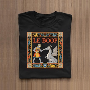 May include: A black t-shirt featuring a medieval-style graphic print with the text LE BOOP. The design shows a knight in chainmail touching the nose of a gray dragon, framed by a colorful border of floral patterns and small animals.