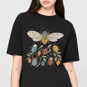 以下が含まれることがあります： 黒いTシャツで、大きなセミのグラフィックプリントが中央にあり、その下には緑の葉がついた枝にとまる様々なカラフルな甲虫や昆虫、そして小さなオレンジと青の種子鞘が描かれています。