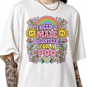 Op de afbeelding: Een gebroken wit t-shirt met een kleurrijke grafische print van een regenboog, zonnebloemen en vlinders. De tekst luidt I NEED 2 MALE VOLUNTEERS FOR A POO in dikke letters. Het ontwerp bevat kleine sterretjes en bloemen.