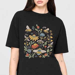 Op de afbeelding: Een zwart T-shirt met korte mouwen en een centrale grafische print van diverse wilde bloemen, meerdere vliegende bijen en kleine rode lieveheersbeestjes. De botanische illustratie bevat gele, oranje en blauwe bloemen met groen blad.