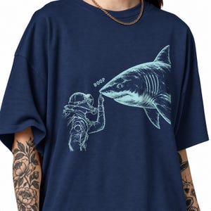 Puede incluir: Una camiseta azul marino con un estampado gráfico en azul claro de un buceador tocando la nariz de un gran tiburón. El texto "BOOP" está impreso en azul claro sobre el dedo del buceador, que se extiende hacia el hocico del tiburón.