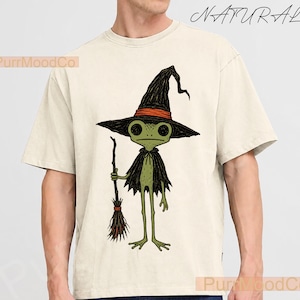 Peut inclure: Un t-shirt à manches courtes de couleur naturelle avec une illustration graphique d'une grenouille verte portant un chapeau de sorcière noir avec une bande orange et une cape noire, tenant un balai marron. Le texte "NATURAL" est en haut à droite.