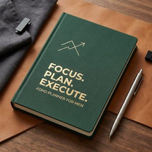 May include: A dark green planner with gold lettering that reads "FOCUS. PLAN. EXECUTE." and "ADHD PLANNER FOR MEN." A silver pen rests beside the planner on a brown leather surface. A gray cloth and a black clip are also visible.