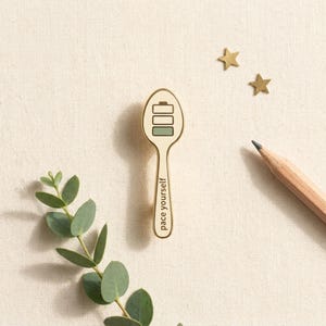 May include: A gold-colored spoon-shaped pin with a battery level indicator and the words "pace yourself." The pin is on a cream-colored surface, with two gold star-shaped embellishments, a pencil, and green foliage.