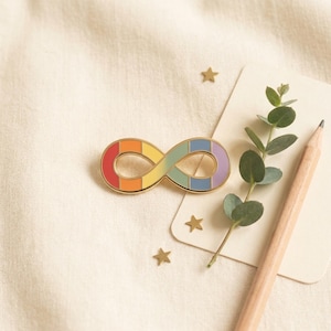 May include: A gold-colored infinity symbol pin with a rainbow gradient design. The pin is on a cream-colored surface, next to a small card, a pencil, and a sprig of greenery. Small gold star-shaped confetti is scattered around.