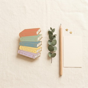 May include: A stack of six colorful book pins with gold trim. The books are in shades of red, green, blue, yellow, and purple. A wooden pencil, sprig of eucalyptus, blank card, and gold star-shaped confetti are also present.