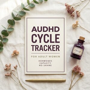 May include: A cream-colored journal with the text "AUDHD CYCLE TRACKER" in dark purple, with the words "FOR ADULT WOMEN" below. A small purple bottle with a "CYCLE" label and a gold pen are also present.
