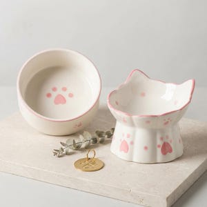 Pode incluir: Duas tigelas brancas de cerâmica para gatos com pegadas e bordas rosa. Uma tigela é redonda, a outra em forma de gato e elevada. Uma etiqueta dourada para animais de estimação com uma pegada também está presente.