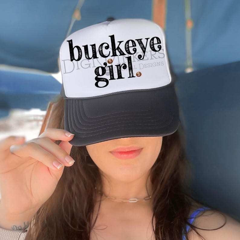 Puede incluir: Gorra de camionero blanca y negra con el texto "buckeye girl" en escritura negra. La gorra tiene una parte trasera de malla y una visera curva. La gorra est&aacute; siendo sostenida por una persona.