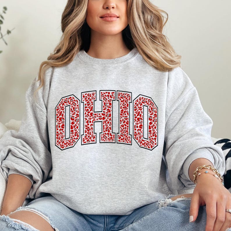 Puede incluir: Sudadera gris claro con la palabra "OHIO" en letras grandes y audaces. Las letras est&aacute;n rellenas con un estampado de animales rojo y blanco y delineadas en negro. Una prenda casual.