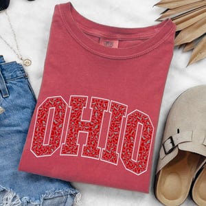 Puede incluir: Una camiseta roja con la palabra "OHIO" en letras grandes con contorno blanco. Las letras est&aacute;n rellenas con un dise&ntilde;o de estampado de leopardo rojo y gris. La camiseta est&aacute; doblada y colocada sobre una superficie blanca junto a unos shorts vaqueros y unas sandalias beige.