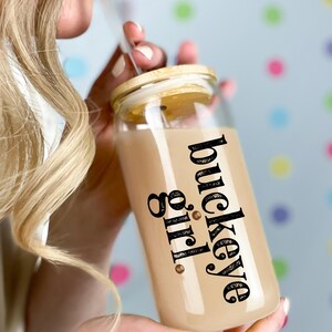 Puede incluir: Un vaso de vidrio transparente con tapa de bamb&uacute; y pajita. El vaso est&aacute; lleno de un l&iacute;quido marr&oacute;n claro y presenta las palabras "buckeye girl" en escritura negra. El fondo es azul claro con lunares de colores.