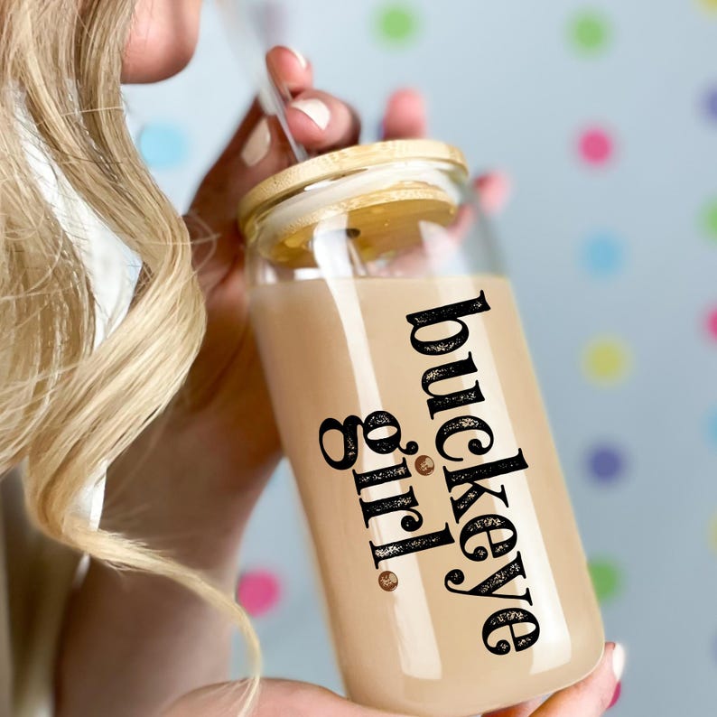 Puede incluir: Un vaso de vidrio transparente con tapa de bamb&uacute; y pajita. El vaso est&aacute; lleno de un l&iacute;quido marr&oacute;n claro y presenta las palabras "buckeye girl" en escritura negra. El fondo es azul claro con lunares de colores.