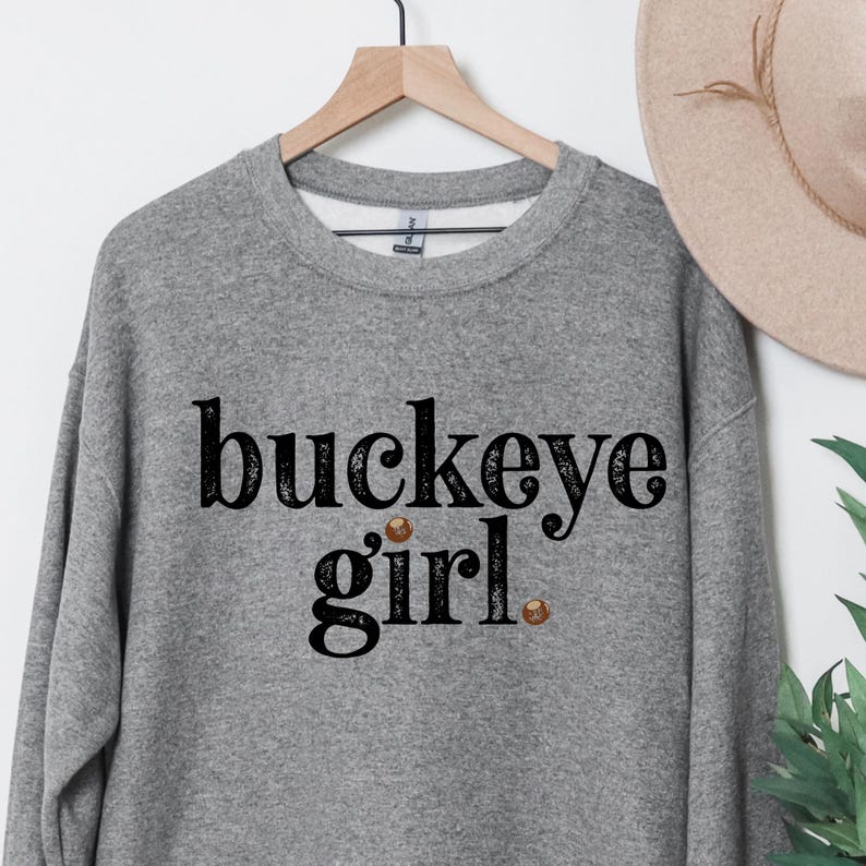 Puede incluir: Una sudadera gris jaspeado con el texto "buckeye girl" en fuente negra. La palabra "buckeye" est&aacute; arqueada sobre la palabra "girl". La sudadera est&aacute; colgada de una percha de madera.