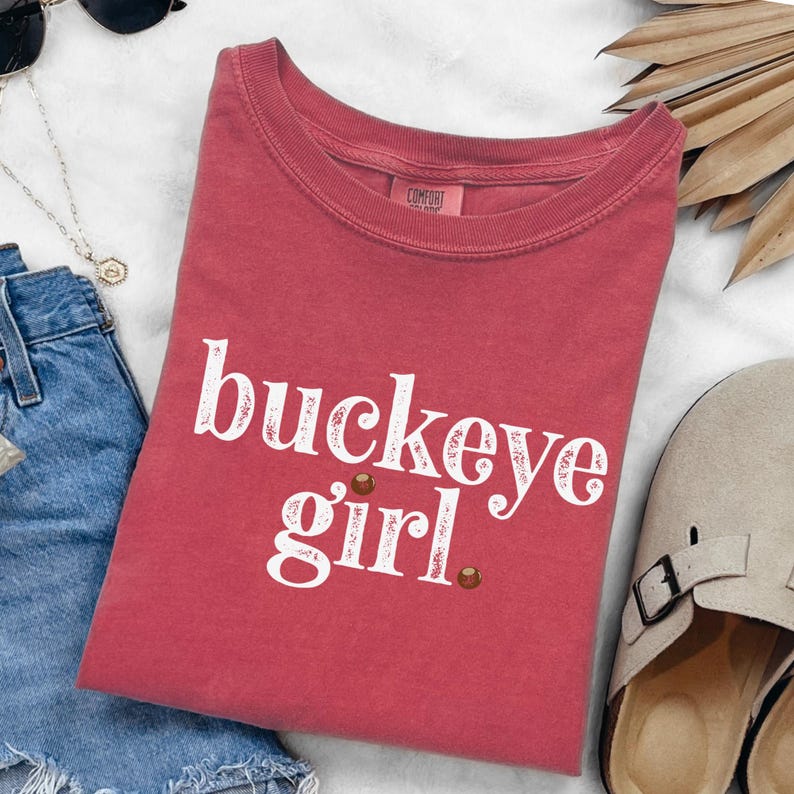 Puede incluir: Una camiseta roja con las palabras "buckeye girl" en escritura blanca. La camiseta tiene cuello redondo y mangas cortas. Dos peque&ntilde;os puntos marrones est&aacute;n debajo de la palabra "girl".