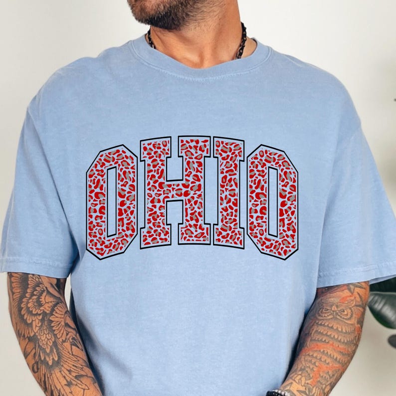Puede incluir: Una camiseta azul claro con la palabra "OHIO" en letras grandes. Las letras est&aacute;n rellenas con un estampado de leopardo rojo y gris, y delineadas en negro.