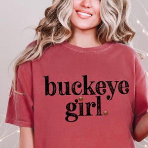 Puede incluir: Camiseta de color granate con la frase "buckeye girl" en negro, con una fuente decorativa. La camiseta tiene cuello redondo y mangas cortas. El texto incluye dos peque&ntilde;os adornos marrones.