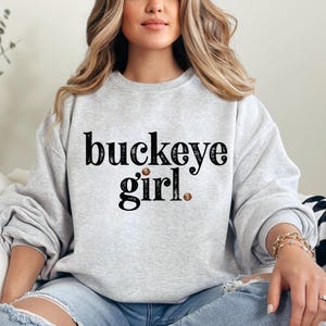 Puede incluir: Sudadera gris claro con la frase "buckeye girl" en negro. La palabra "buckeye" es m&aacute;s grande que "girl". Dos peque&ntilde;os puntos marrones forman parte del dise&ntilde;o. La sudadera es de cuello redondo.