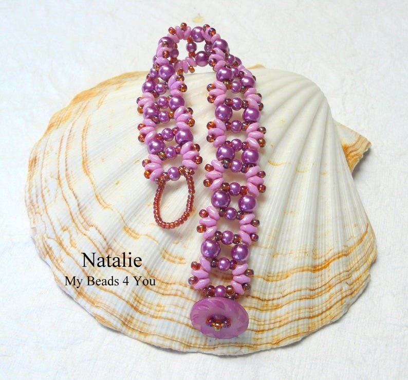 K&ouml;nnte beinhalten: Ein rosa und violettes Perlenarmband mit Blumenmuster. Das Armband liegt auf einem wei&szlig;en Muschelhintergrund. Der Text "Natalie My Beads 4 You" ist unterhalb des Armbands sichtbar.