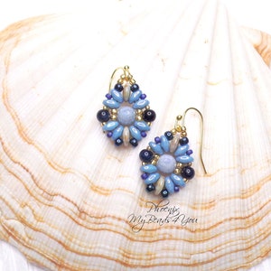 May include: A pair of drop earrings featuring a central light blue bead surrounded by a floral pattern of blue, dark blue, and metallic gold seed beads. The earrings are suspended from gold-toned metal hooks and include the text Phoenix MyBeads4You.