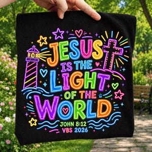 May include: A black fabric item featuring a colorful neon-style graphic with the text "JESUS IS THE LIGHT OF THE WORLD" and "JOHN 8:12 VBS 2026". The design includes a lighthouse, a cross, stars, and hearts in vibrant shades of pink, blue, yellow, and green.