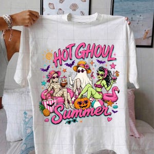Puede incluir: Una camiseta blanca con un gr&aacute;fico colorido y el texto "HOT GHOUL Summer". El dise&ntilde;o muestra una momia, un fantasma y un monstruo verde con gafas de sol y trajes de ba&ntilde;o, rodeados de art&iacute;culos de playa como un flotador de flamenco y una calabaza.