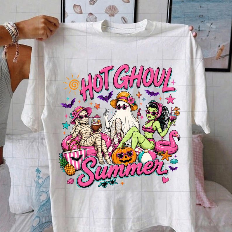 Puede incluir: Una camiseta blanca con un gr&aacute;fico colorido y el texto "HOT GHOUL Summer". El dise&ntilde;o muestra una momia, un fantasma y un monstruo verde con gafas de sol y trajes de ba&ntilde;o, rodeados de art&iacute;culos de playa como un flotador de flamenco y una calabaza.