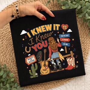 K&ouml;nnte beinhalten: Ein schwarzes T-Shirt mit einem Grafikdruck mit dem Text I KNEW IT I KNEW YOU, INFINITY AND BEYOND und YOU'VE GOT A FRIEND IN ME. Das Design zeigt eine Person mit langem braunem Haar, eine Gitarre, einen Kaktus und Cowboystiefel.