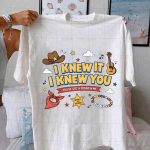 K&ouml;nnte beinhalten: Ein wei&szlig;es Kurzarm-T-Shirt mit einem Grafikdruck, der einen braunen Cowboyhut, eine Gitarre, ein rotes Bandana, einen Sheriffstern mit der Aufschrift Infinity and Beyond und ein Perlenarmband zeigt. Der Text lautet I KNEW IT I KNEW YOU YOU'VE GOT A FRIEND IN ME.