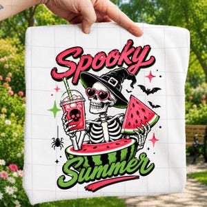 Puede incluir: Una toalla blanca con un gr&aacute;fico de un esqueleto que lleva un sombrero de bruja negro y gafas de sol rosas en forma de coraz&oacute;n. El esqueleto sostiene una bebida rosa con un icono de calavera y una rodaja de sand&iacute;a, sentado dentro de una sand&iacute;a.