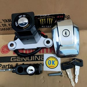 May include: A collection of motorcycle ignition components, including a black and silver ignition switch, a chrome fuel tank cap, a black lock, two keys, and a bolt. The ignition switch has "OFF ON" and "IGNITION" labels. A "QUALITY INSPECTION OK YAMAHA" sticker is also present.